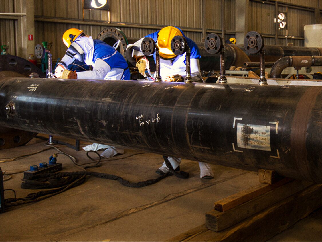 Two welders in hard hats working on a large-bore pipe spool in a fabrication shop